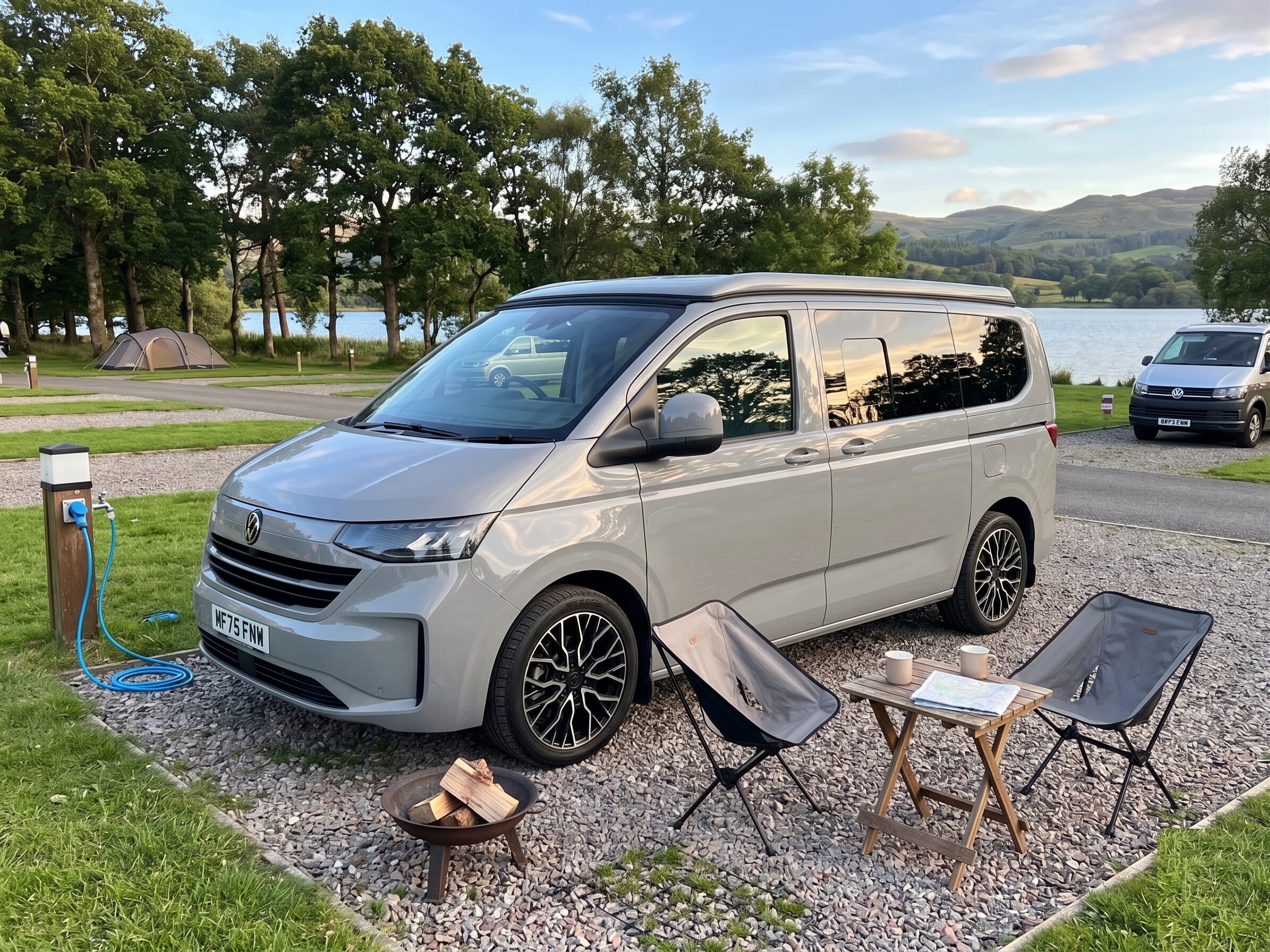 Grey VW Transporter campervan pitched by a lake with camping chairs and a fire pit